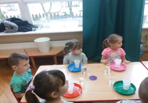 dzieci siedzą przy stolikach a na talerzach kolorowe kubki ze słomką